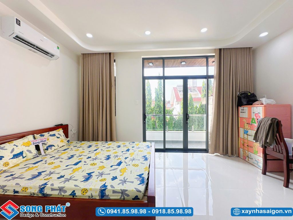 Không gian phòng ngủ rộng rãi, bố trí giường ngủ gỗ, rèm cửa che kín ánh sáng và hệ tủ kệ gọn gàng bên góc phòng.