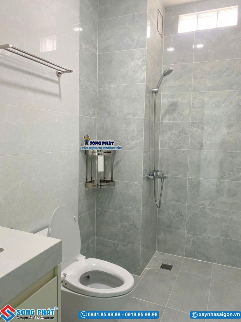 Khu vực phòng tắm và vệ sinh lát gạch xám vân đá sạch sẽ, trang bị vòi sen đứng.
