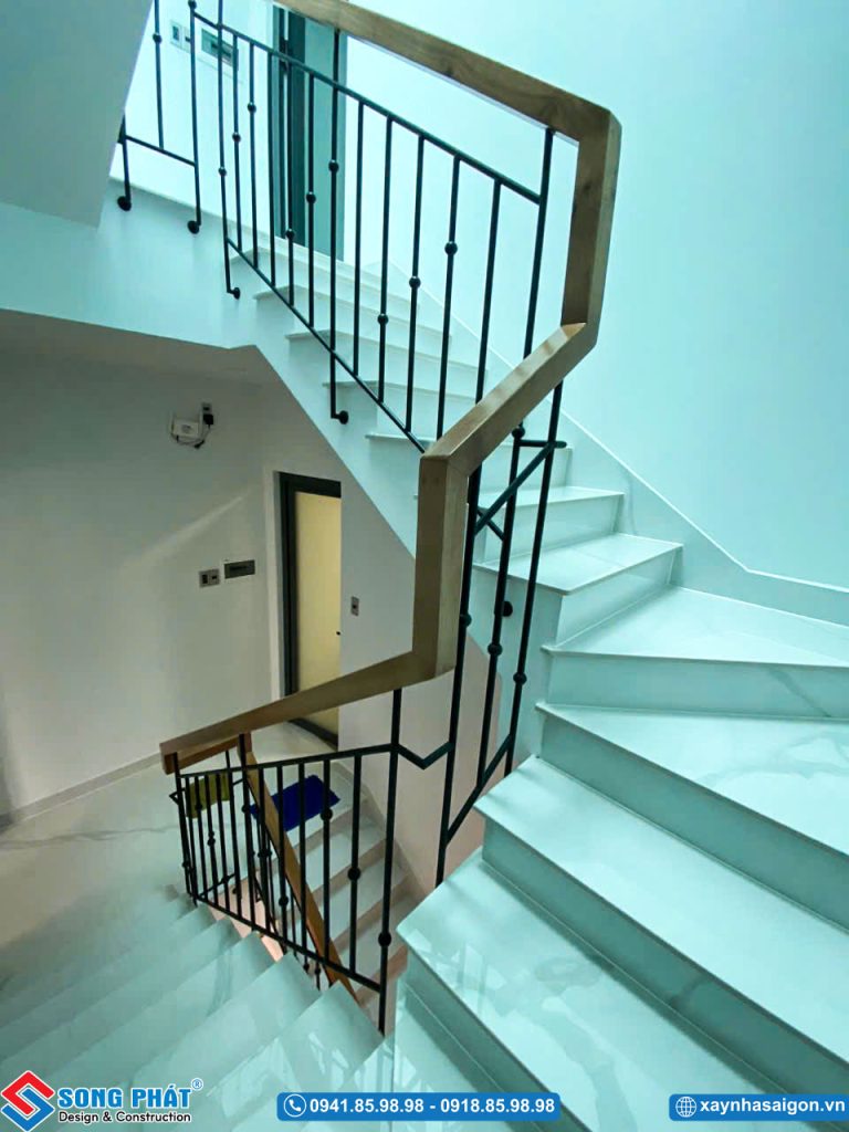 Cầu thang uốn lượn lát đá trắng, kết hợp lan can sắt tay vịn gỗ chắc chắn.